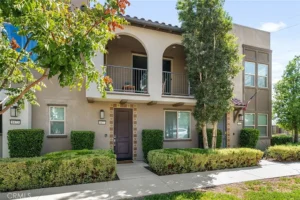 Two-story Southern California townhouse with balcony, arched openings, dark brown door, and manicured bushes and trees.