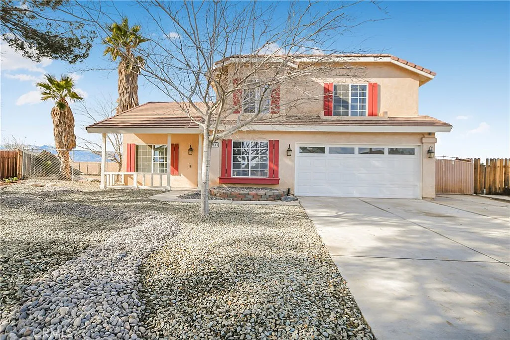 Two-story beige house with red shutters, stone landscaping, palm trees, and a white garage.