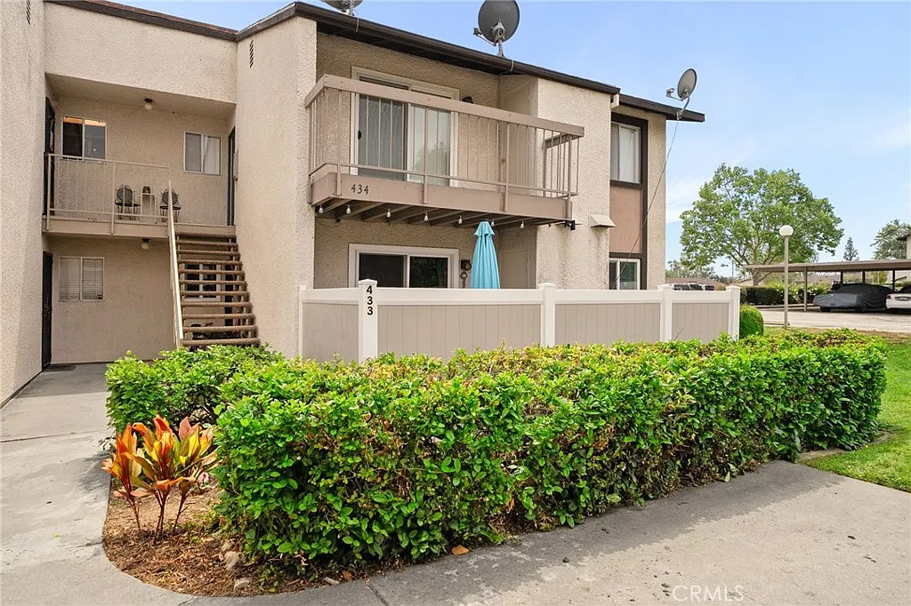 Two-story beige apartment building with balcony, numbered railing, hedge, plants, and fenced patio.