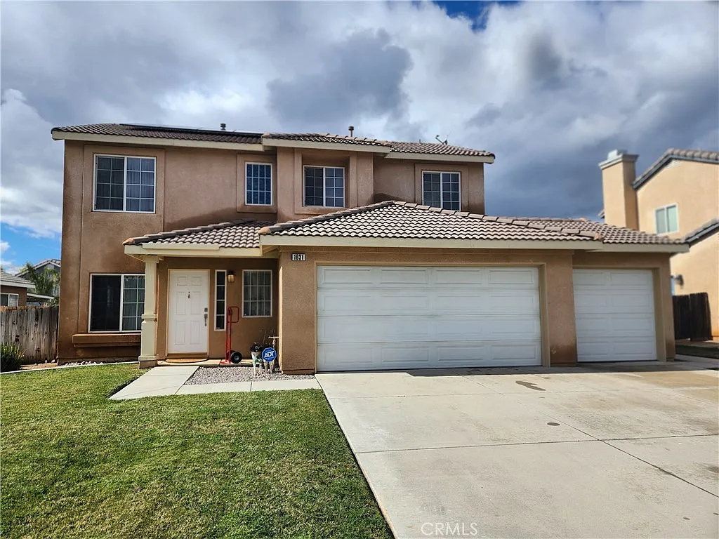 Two-story beige Southern California house with three-car garage, tiled roof, white windows, and a trimmed lawn.