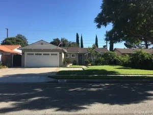 Front view of a Southern California gray house with two-car garage, manicured lawn, American flag, and tall trees.