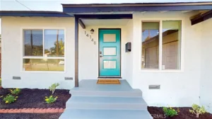 Light blue front door with glass panels and house number 14148 on a white Southern California house, flanked by windows, welcome mat, and small plants.