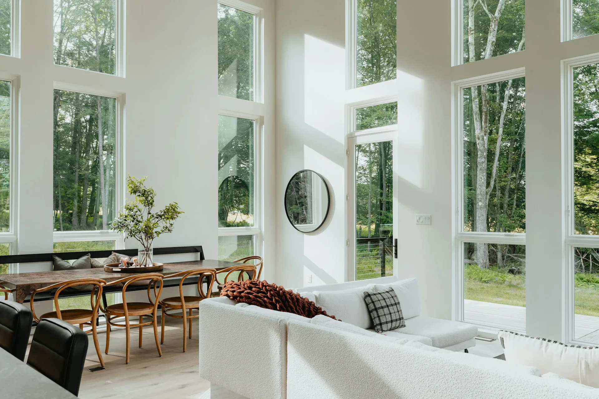 Modern living room in California with tall windows, forest view, and light pouring in. Features a cozy white sofa, round mirror, and a wooden dining set.